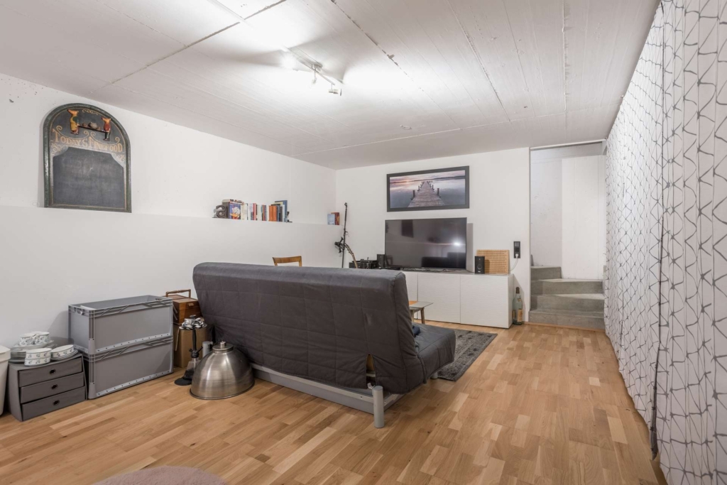 Ein moderner Kellerraum mit hellem Holzfußboden, einem grauen Futon, einem Fernseher, kleinen Regalen, einem Wandspiegel und einer minimalistischen Einrichtung. Eine Treppe führt auf eine höhere Ebene, und ein großer gemusterter Vorhang bedeckt eine Wand.