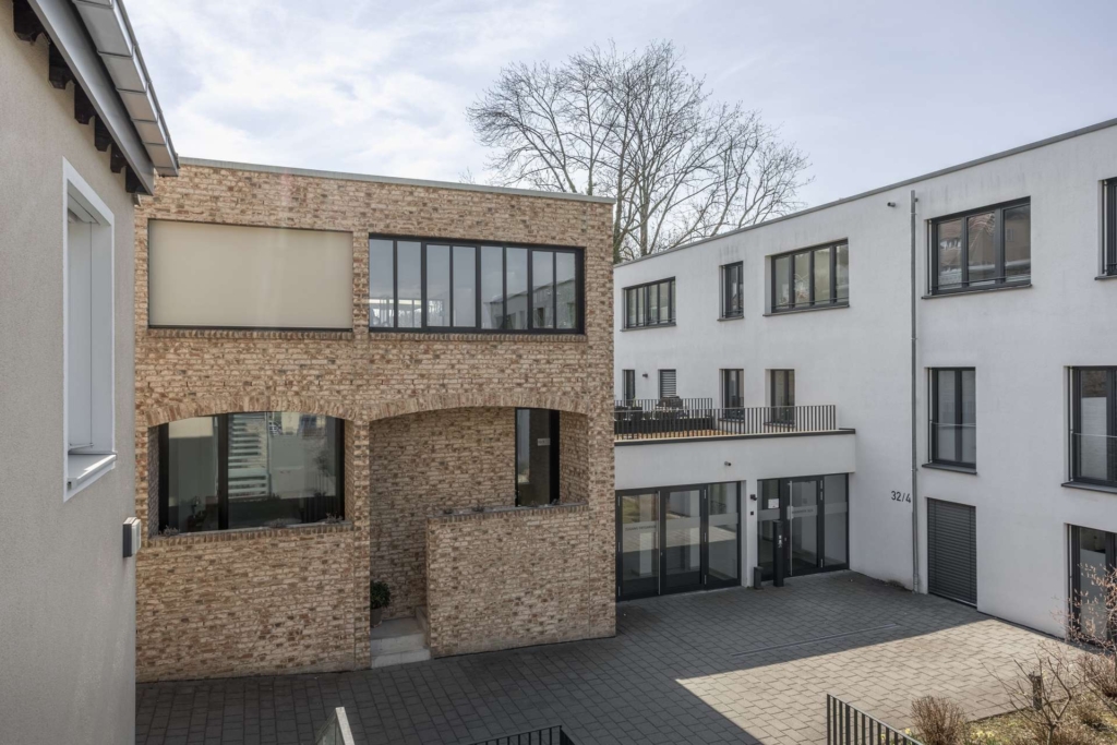Moderne Wohngebäude mit großen Fenstern und einem zentralen Steinbau, die an einem sonnigen Tag einen gepflasterten Innenhof umgeben. Ein blattloser Baum erhebt sich hinter den Gebäuden.