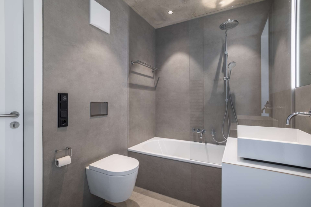 Modernes Badezimmer mit grauen Wänden, einer wandmontierten Toilette, einer Badewanne mit Duschkopf, einem weißen Waschbecken auf einem Waschtisch, einem großen Spiegel und minimalistischen Armaturen. Der Raum ist sauber und aufgeräumt mit zeitgenössischem Design.