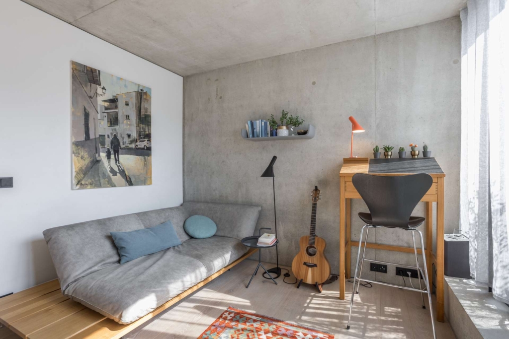 Ein modernes, minimalistisches Wohnzimmer mit einem grauen Sofa, einem Schreibtisch mit einer orangefarbenen Lampe und einem schwarzen Stuhl, einer Gitarre, einer Stehlampe, Wandkunst, einem schwebenden Bücherregal, Topfpflanzen und großen Fenstern, die natürliches Licht hereinlassen.