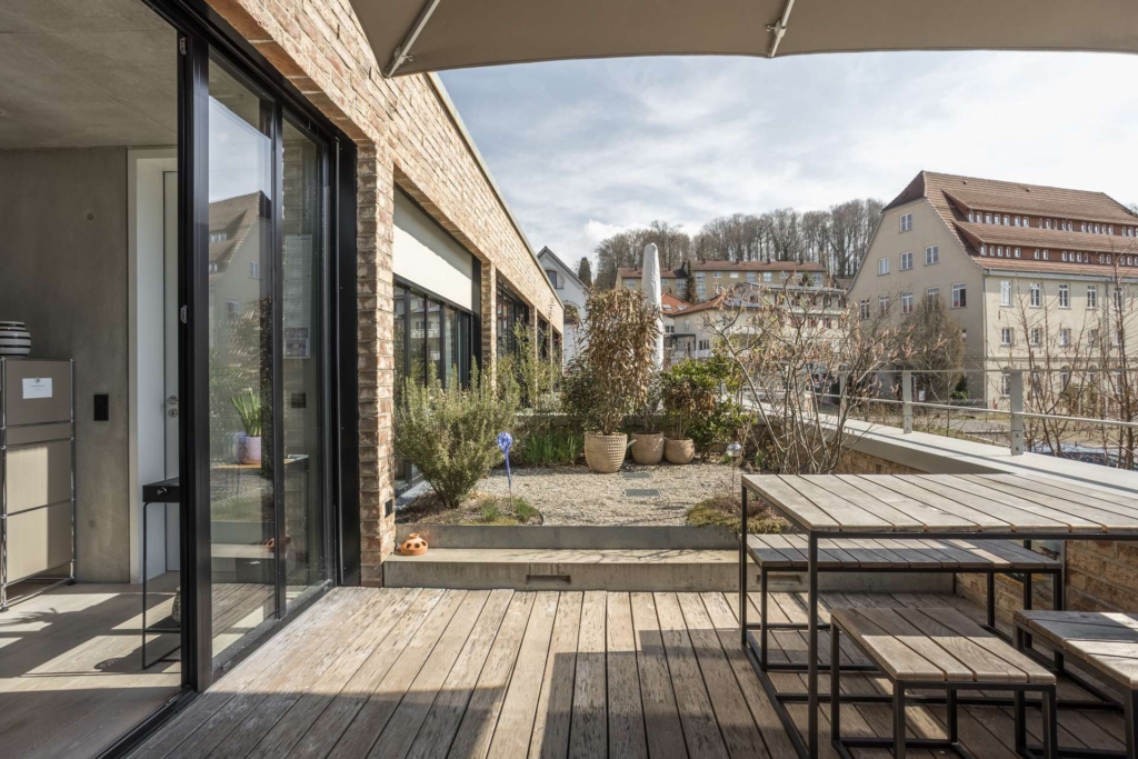 Eine moderne Dachterrasse mit Holzbelag, Metalltischen und -bänken, Topfpflanzen und Hochbeeten. Die umliegenden Gebäude und Bäume sind bei teilweise bewölktem Himmel zu sehen.