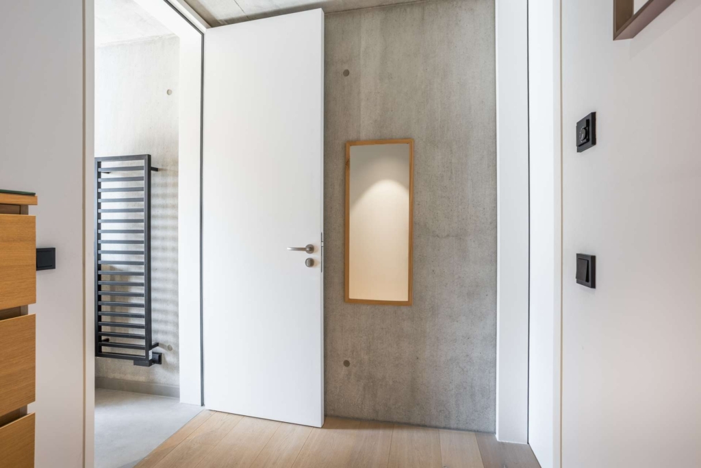 Moderner minimalistischer Flur mit einer teilweise offenen weißen Tür, einem kleinen Spiegel mit Holzrahmen an einer Betonwand und einem schwarzen Handtuchhalter, der in einem angrenzenden Badezimmer zu sehen ist. Helle Holzböden verleihen dem Raum Wärme.