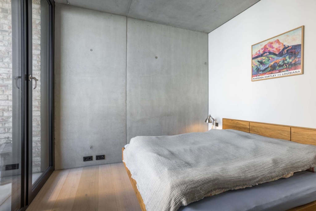 Ein minimalistisches Schlafzimmer mit einer Betonwand, einem Holzboden, einer großen Glastür, einem Holzbett mit einer grauen Decke, einer kleinen Nachttischlampe und einem bunten Gemälde an der weißen Wand.