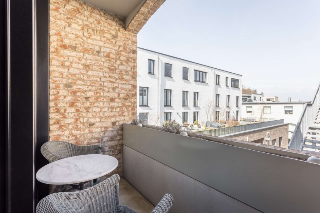 Ein kleiner Balkon mit zwei Korbsesseln und einem runden Marmortisch bietet an klaren Tagen einen Blick auf moderne weiße Wohnhäuser. Auf einer Seite des Balkons befindet sich eine Backsteinmauer.