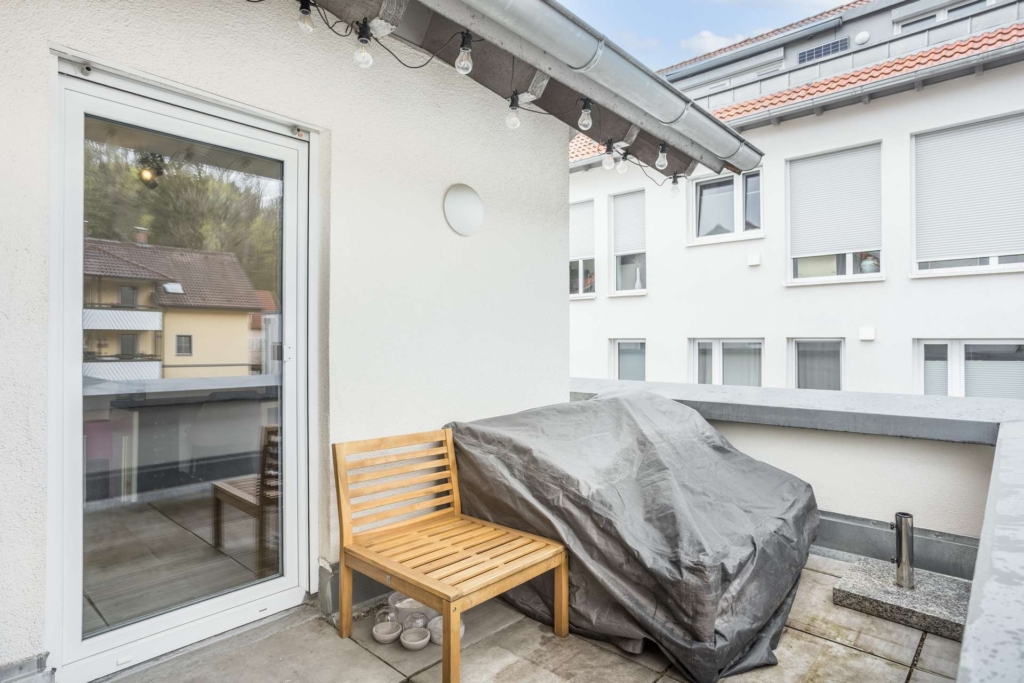 Ein kleiner Balkon mit einer Holzbank, einem Grill, der mit einer grauen Plane abgedeckt ist, und einer Glasschiebetür. Unter dem Dach hängen Lichterketten, und auf der anderen Straßenseite sind Gebäude mit geschlossenen Fensterläden zu sehen.