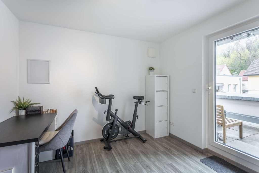 Ein modernes, minimalistisches Heimbüro mit einem Schreibtisch, einem grauen Stuhl, einer Zimmerpflanze, Büchern, einem Heimtrainer, einem weißen Schrank und einem großen Fenster, das auf einen Balkon mit Sitzgelegenheiten im Freien und Blick auf Häuser und Bäume führt.