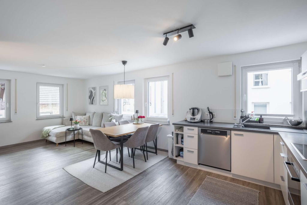 Ein moderner, offener Küchen- und Essbereich mit hellen Holzböden, weißen Wänden und großen Fenstern. Der Raum umfasst einen Esstisch mit Stühlen, ein graues Sofa und moderne Küchengeräte.