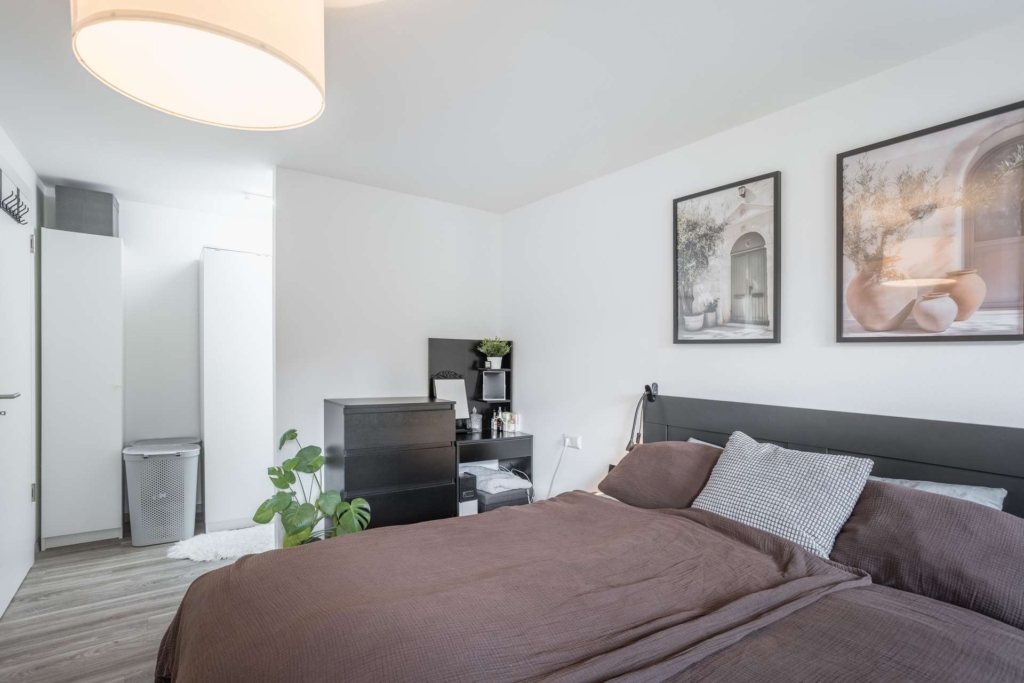 Ein modernes Schlafzimmer mit einem ordentlich gemachten Bett, braunem Bettzeug, schwarzem Kopfteil, schwarzer Kommode, Topfpflanze, Wandkunst und hellem Holzfußboden. Weiße Wände und sanfte Beleuchtung schaffen eine saubere, gemütliche Atmosphäre.
