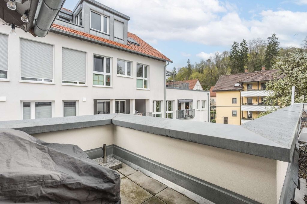Blick von einem kleinen Balkon mit überdachtem Grill auf moderne Wohngebäude und Bäume unter einem teilweise bewölkten Himmel.