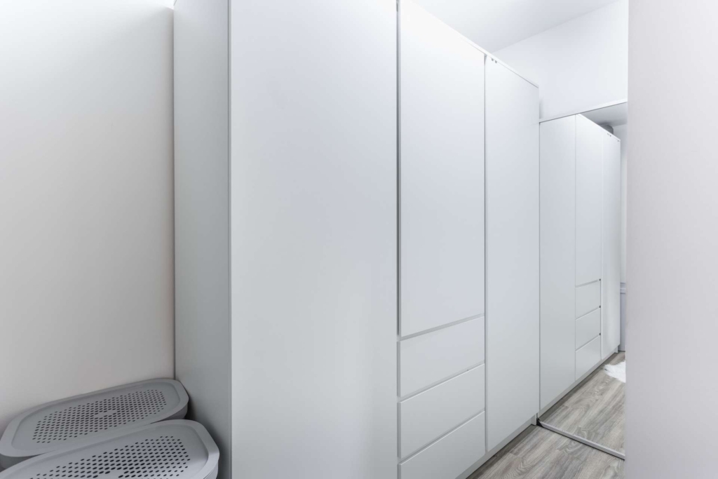 Ein moderner, minimalistischer Kleiderschrank mit weißen Schränken und eingebauten Schubladen, einer verspiegelten Schiebetür, hellem Holzboden und zwei grauen Wäschekörben in der Ecke. Der Raum ist hell und sauber.