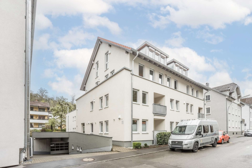 In einer ruhigen Straße steht ein modernes, weißes, dreistöckiges Wohnhaus mit Balkonen und einem Schrägdach. Ein Lieferwagen und ein orangefarbenes Auto sind in der Nähe geparkt, und eine Einfahrt zu einer Tiefgarage ist zu sehen. Der Himmel ist teilweise bewölkt.