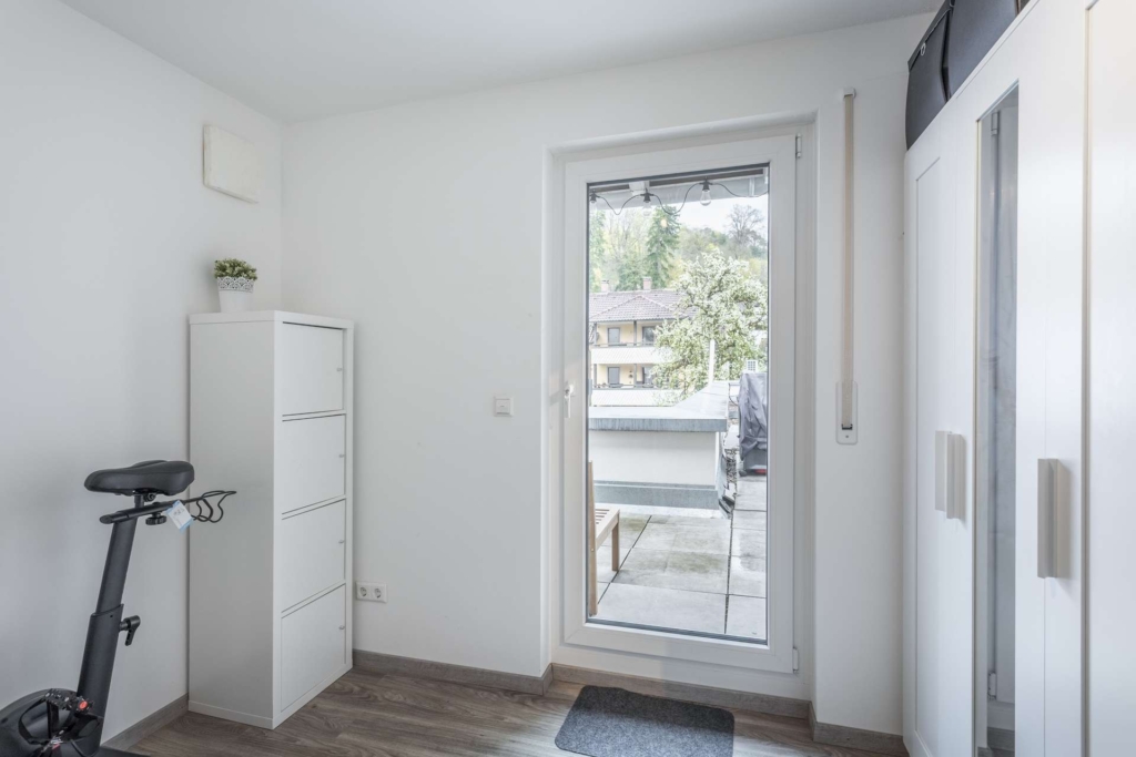Ein helles Zimmer mit weißen Wänden, einem Heimtrainer, einem hohen weißen Schrank und einem Kleiderschrank. Eine Glastür führt auf einen Balkon mit Gartenmöbeln und Blick auf Grünflächen und Gebäude.