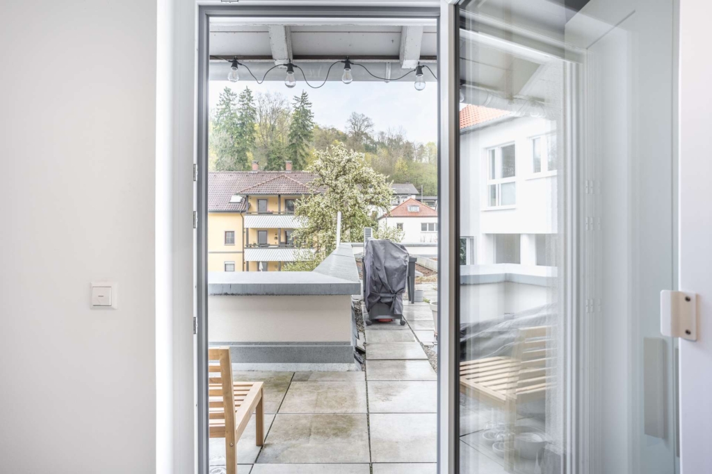 Blick aus einem modernen Innenraum durch eine Glastür auf einen Balkon mit einem Grill, Holzstühlen und Wohngebäuden, Bäumen und einem bewölkten Himmel im Hintergrund.