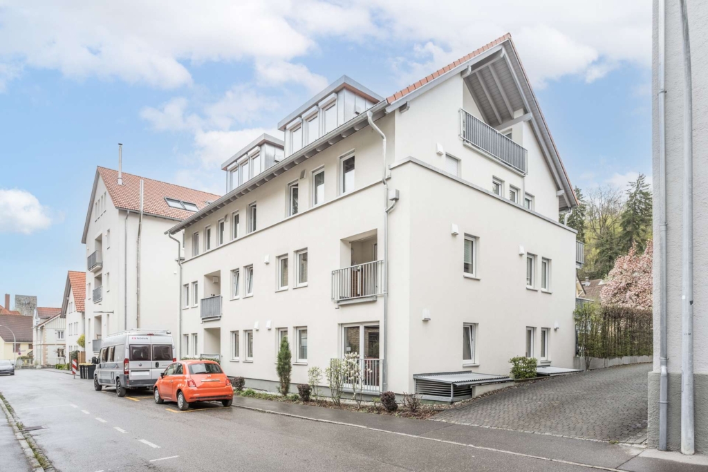 Ein modernes, weißes, dreistöckiges Wohnhaus mit Balkonen und Schrägdach in einer ruhigen Straße mit geparkten Autos und wolkenverhangenem Himmel über uns.