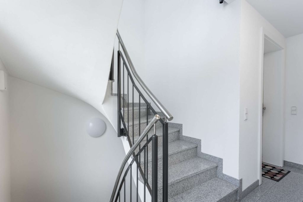 Eine moderne Innentreppe mit grauen Stufen und einem schwarzen Metallgeländer, die sich neben einer weißen Wand nach oben wölbt. Links ist eine runde Wandleuchte angebracht, rechts befindet sich eine offene Tür mit einer gemusterten Matte.