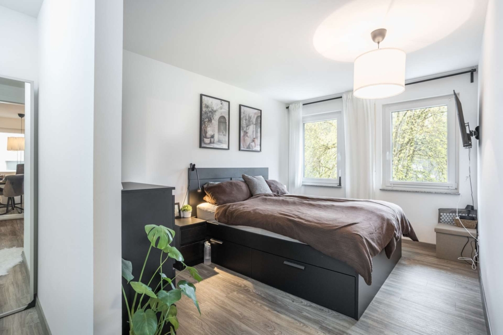 Modernes Schlafzimmer mit einem großen Bett aus dunklem Holz, braunem Bettzeug, zwei Fenstern mit Bäumen draußen, Wandkunst über dem Bett, einem Nachttisch mit einer Pflanze und Holzfußböden. Helle, minimalistische Einrichtung mit natürlichem Licht.