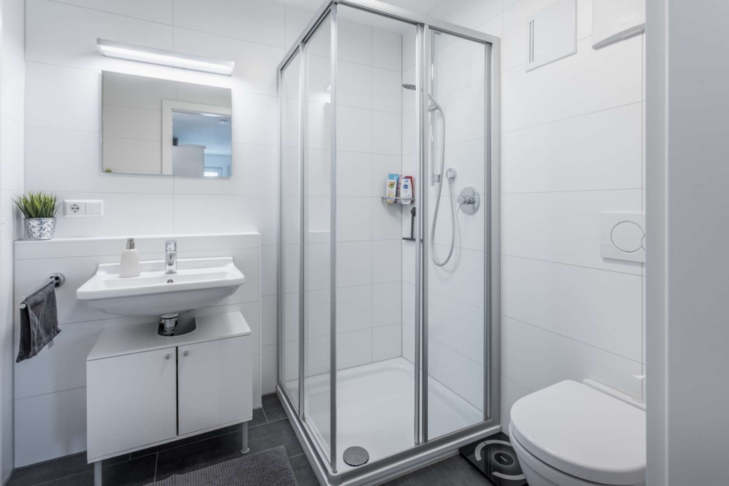 Modernes Badezimmer mit weißen Wänden, Glasduschabtrennung, weißem Waschbecken mit Unterschrank, Spiegel, Seifenspender, kleiner Topfpflanze, Handtuchaufhängung, wandmontierter Toilette und grauem Fußbodenbelag.