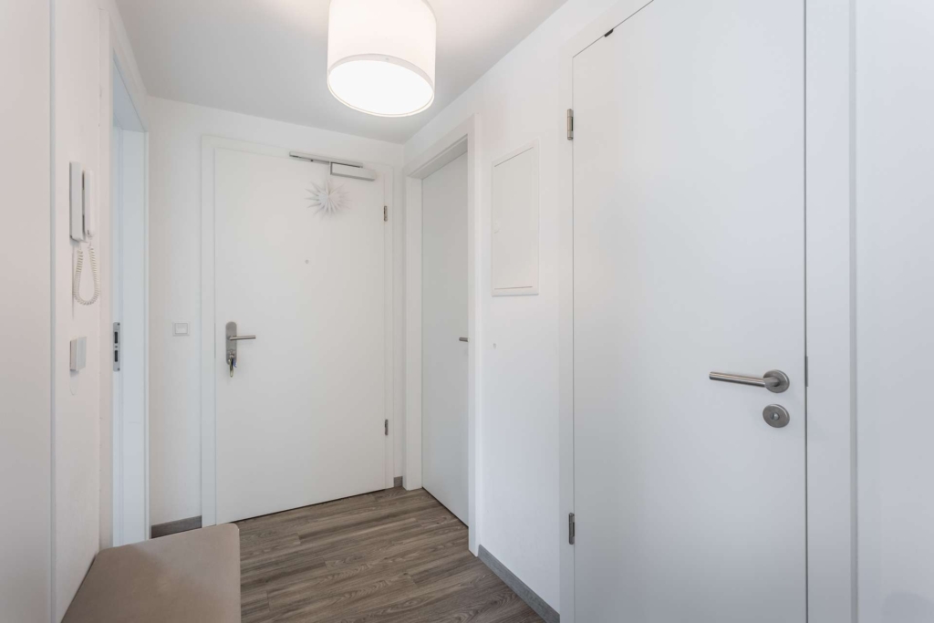 Ein heller, minimalistischer Flur mit weißen Wänden und Türen, einer Deckenleuchte, Holzböden und einer Gegensprechanlage an der Wand.