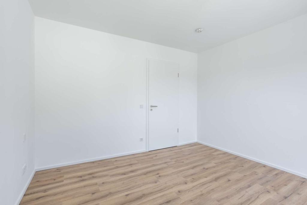 Ein leerer Raum mit weißen Wänden, einer geschlossenen weißen Tür und einem hellen Holzfußboden. Der Raum ist durch natürliches Licht gut beleuchtet und wirkt dadurch sauber und minimalistisch.