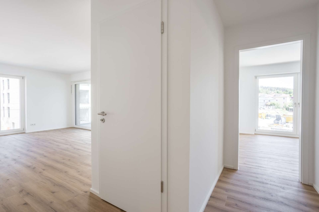 Helle, leere Wohnung mit weißen Wänden, hellen Holzböden und großen Fenstern. Die Ansicht zeigt einen Flur mit offenen Türen, die zu zwei geräumigen, sonnendurchfluteten Zimmern führen.