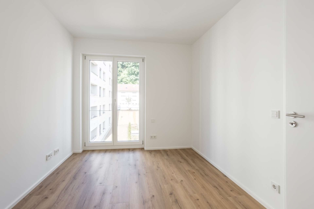 Ein heller, leerer Raum mit weißen Wänden, Holzfußboden und einer großen Glastür, die auf einen Balkon führt. Natürliches Licht erfüllt den Raum, und draußen ist ein modernes Gebäude zu sehen.