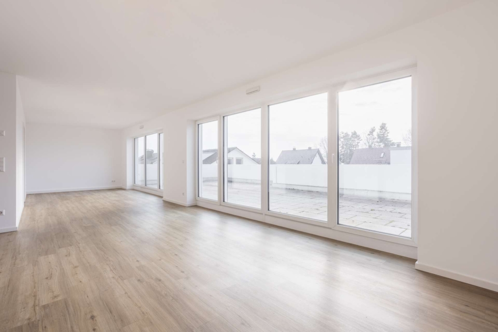 Heller, leerer Raum mit großen, bodentiefen Fenstern, hellem Holzfußboden und weißen Wänden. Die Fenster blicken auf eine geräumige Außenterrasse mit Häusern und Bäumen im Hintergrund.