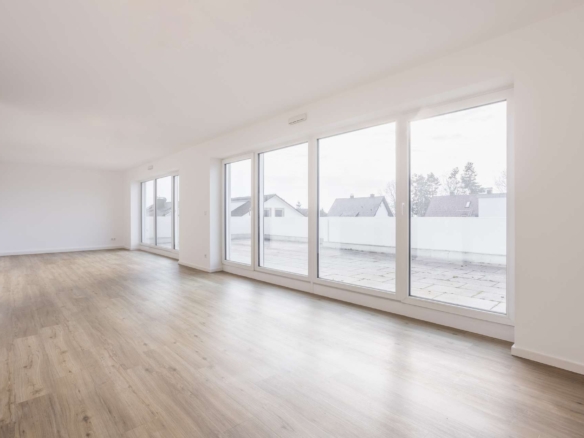 Heller, leerer Raum mit großen, bodentiefen Fenstern, hellem Holzfußboden und weißen Wänden. Die Fenster blicken auf eine geräumige Außenterrasse mit Häusern und Bäumen im Hintergrund.