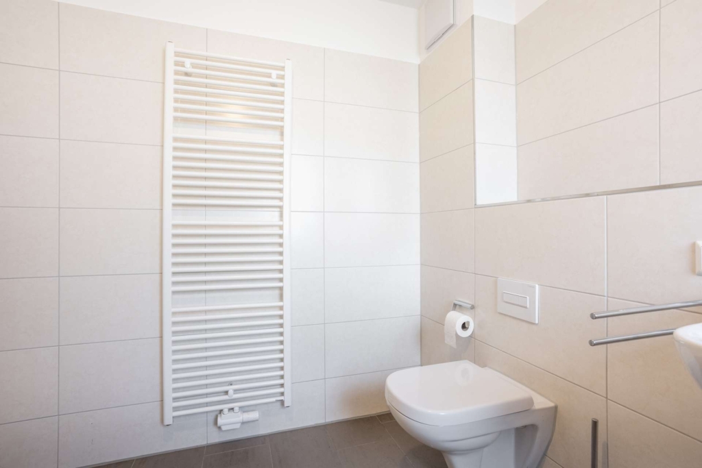 Ein modernes Badezimmer mit beige gefliesten Wänden, einer wandmontierten weißen Toilette, einem Toilettenpapierhalter, einem kleinen Waschbecken und einem hohen weißen Handtuchheizkörper. Der Raum ist minimalistisch und gut beleuchtet.