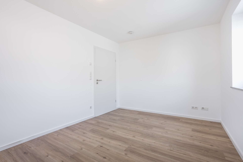 Ein kleiner, leerer Raum mit weißen Wänden, einer geschlossenen weißen Tür, hellem Holzfußboden und einem Fenster auf der rechten Seite, das natürliches Licht hereinlässt. An der unteren rechten Wand befinden sich Steckdosen.