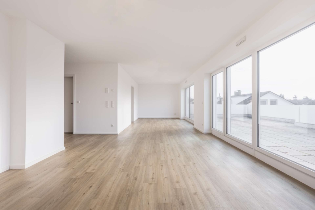 Ein geräumiger, leerer Raum mit hellem Holzfußboden, weißen Wänden und großen, bodentiefen Fenstern, die viel natürliches Licht und einen Blick auf die Terrasse nach draußen bieten.