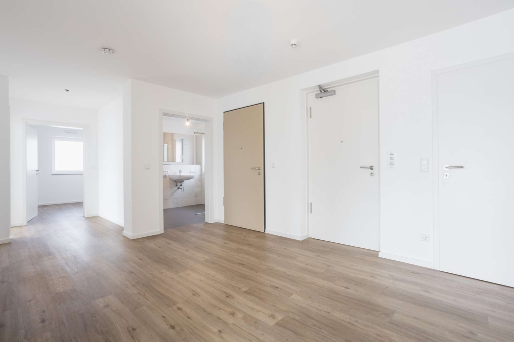 Eine geräumige, leere Wohnung mit weißen Wänden, hellem Holzfußboden, mehreren Türen und einem Blick in ein Badezimmer und ein weiteres helles Zimmer mit Fenster.