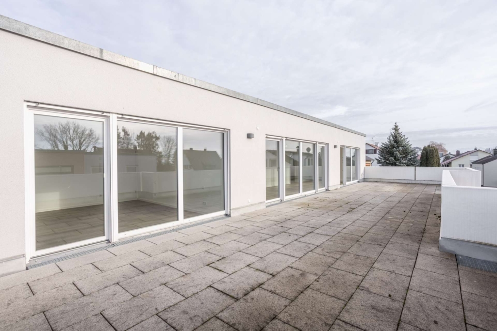 Geräumige Dachterrasse mit hellen Fliesen und einem weißen Geländer, angrenzend an ein modernes Gebäude mit großen Glasschiebetüren und Fenstern. Im Hintergrund sind Bäume und benachbarte Häuser bei bewölktem Himmel zu sehen.