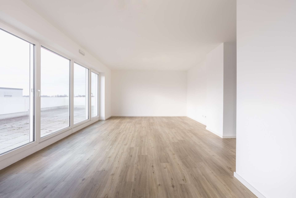 Ein geräumiger, leerer Raum mit hellem Holzfußboden, großen, bodentiefen Fenstern auf der linken Seite und weißen Wänden, die viel natürliches Licht von einer Außenterrasse hereinlassen.