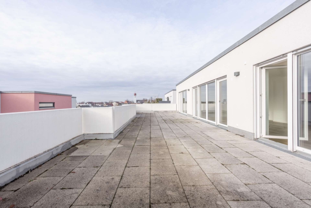 Eine große Dachterrasse mit hellgrauen Fliesen, weißen Wänden und großen Glastüren, die ins Innere führen. Der Himmel ist bedeckt, und in der Ferne sind die Nachbargebäude zu sehen.