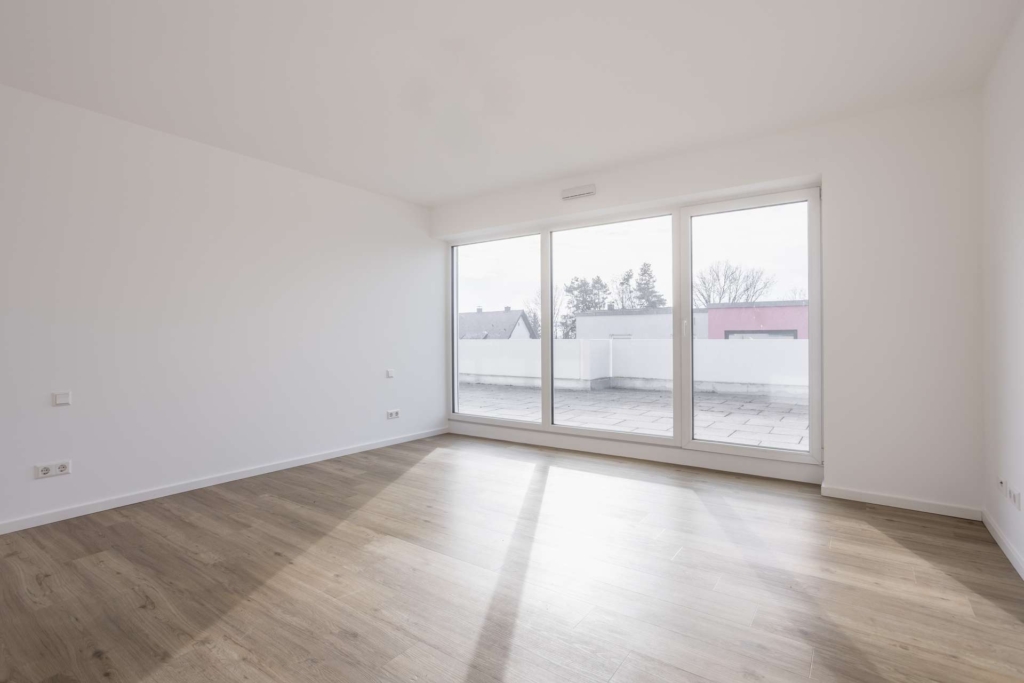 Heller, leerer Raum mit weißen Wänden, hellem Holzfußboden und großen, bodentiefen Fenstern, die auf einen geräumigen Balkon mit Blick auf Bäume und Nachbargebäude führen.