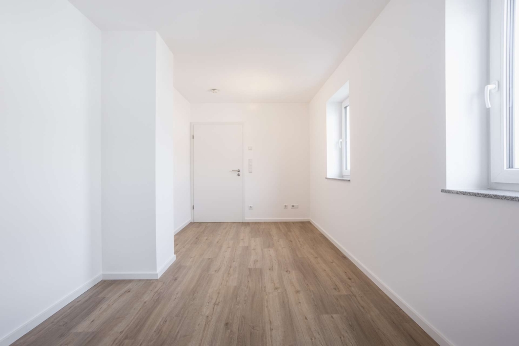 Ein minimalistischer, leerer Raum mit weißen Wänden, einer geschlossenen weißen Tür, zwei Fenstern auf der rechten Seite und hellem Holzfußboden. Das natürliche Licht erhellt den Raum und schafft eine klare und moderne Atmosphäre.