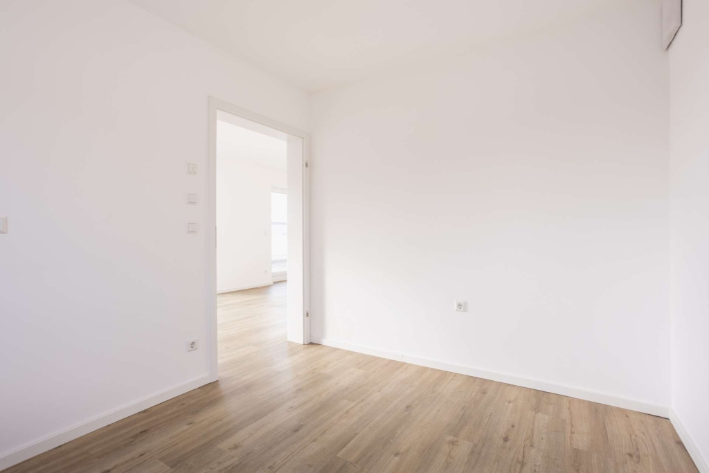 Ein leerer, heller Raum mit weißen Wänden, einem hellen Holzfußboden und einer offenen Tür, die zu einem weiteren sonnendurchfluteten Raum führt. Der Raum hat eine minimale Ausstattung und ein sauberes, modernes Aussehen.