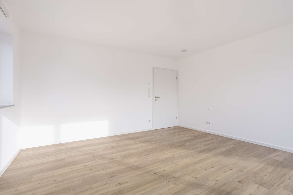 Ein heller, leerer Raum mit weißen Wänden, einer geschlossenen weißen Tür und einem hellen Holzfußboden. Durch ein Fenster strömt Sonnenlicht herein und schafft eine helle und saubere Atmosphäre.