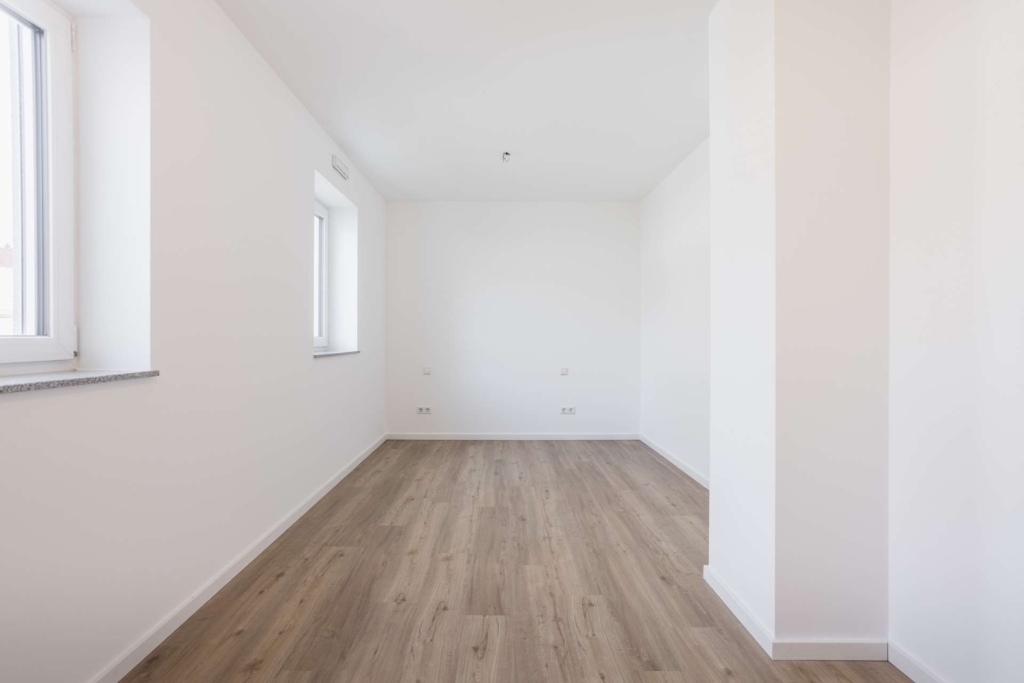 Ein heller, leerer Raum mit weißen Wänden, zwei Fenstern auf der linken Seite und hellem Holzfußboden. Der Raum wirkt frisch renoviert, und natürliches Licht erhellt den minimalistischen Bereich.