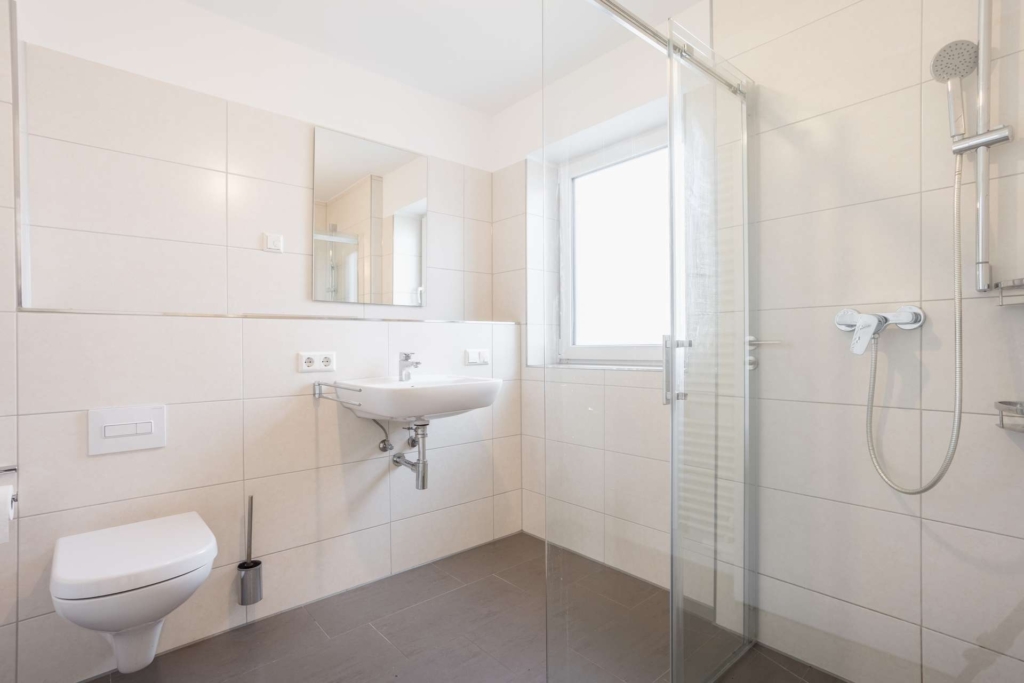 Modernes Badezimmer mit beige gefliesten Wänden, einer an der Wand befestigten Toilette und einem Waschbecken, einem Spiegel über dem Waschbecken, einer verglasten Dusche mit Regendusche und einem großen Fenster, das Tageslicht hereinlässt.