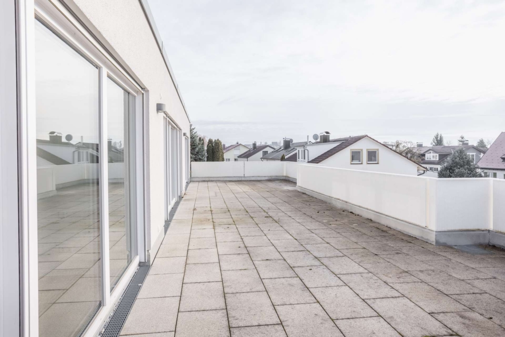 Eine geräumige, leere Dachterrasse mit hellen Fliesen und weißem Geländer, die an ein modernes Gebäude mit großen Glastüren angrenzt, bietet an einem bewölkten Tag einen Blick auf Wohnhäuser.