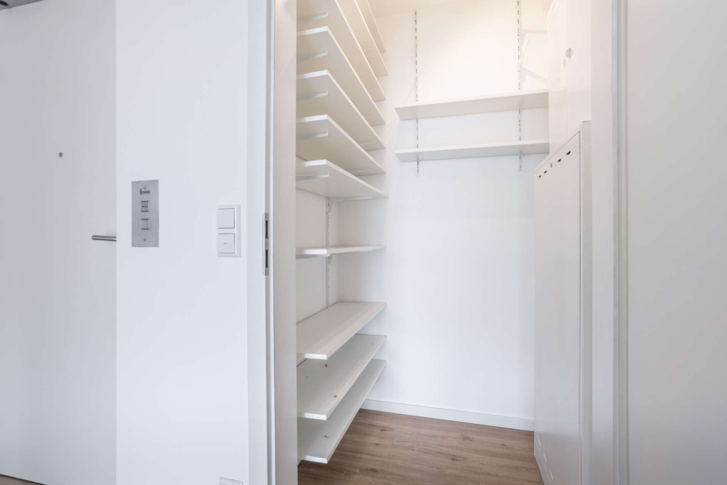 Ein moderner, leerer begehbarer Kleiderschrank mit weißen Regalen und Hängestangen, weißen Wänden und einem hellen Holzboden. Der Schrank ist gut beleuchtet und mit verstellbaren Regalen auf einer Seite organisiert.