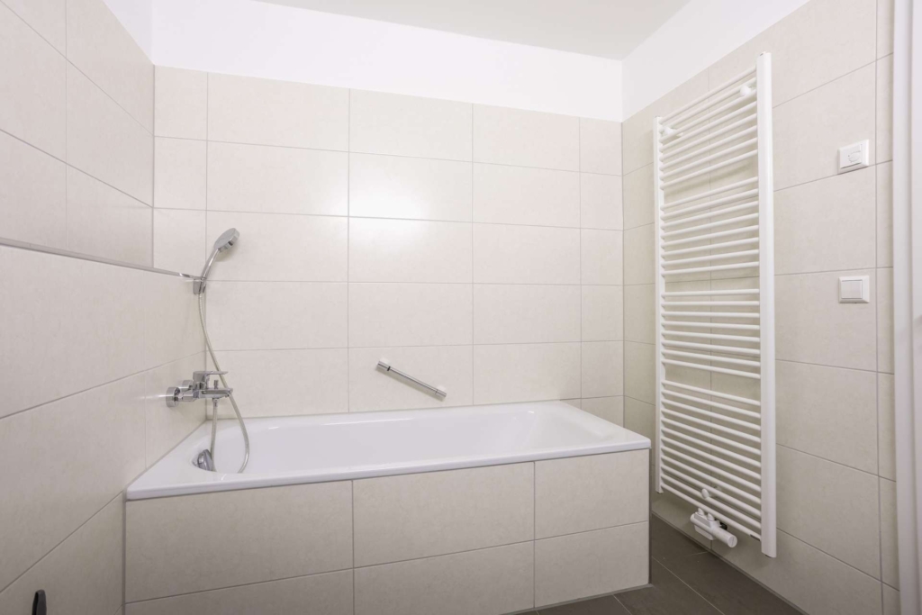 Ein modernes Badezimmer mit beige gefliesten Wänden, einer weißen Badewanne mit silbernem Wasserhahn und Handbrause, einem Haltegriff und einem hohen weißen beheizten Handtuchhalter an der Wand.
