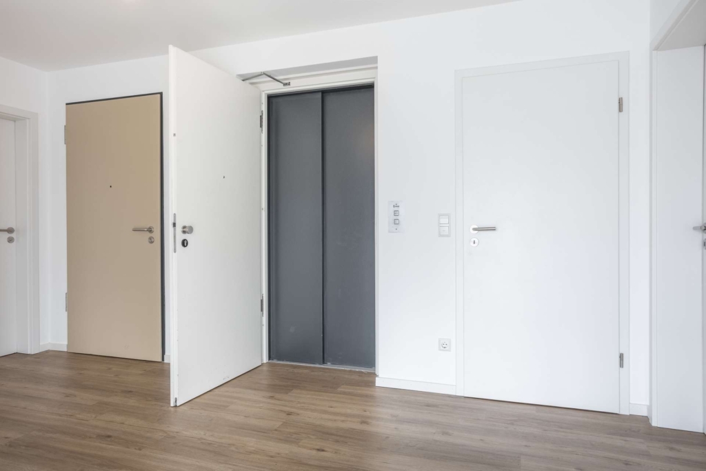 Ein moderner Flur mit hellem Holzfußboden, drei weißen Türen und einem offenen Aufzug mit grauen Türen. Die Wände sind weiß, und der Raum ist hell mit natürlichem Licht.