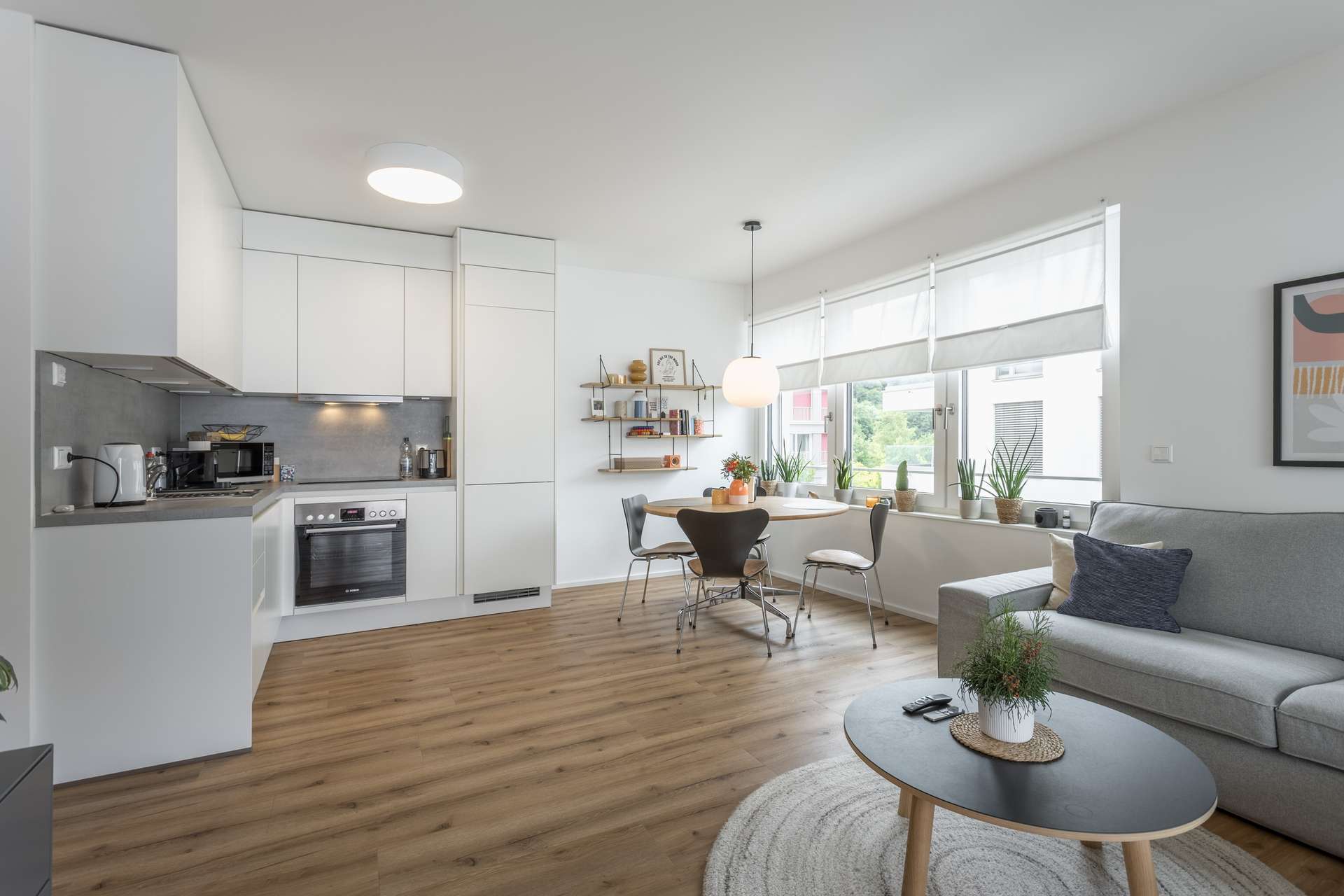Wohn- und Essbereich Moderne, helle, offene Küche und Wohnzimmer mit Holzfußboden, weißen Schränken, einem grauen Sofa, einem runden Couchtisch und einem Essbereich an großen Fenstern mit weißen Jalousien, die viel natürliches Licht hereinlassen.