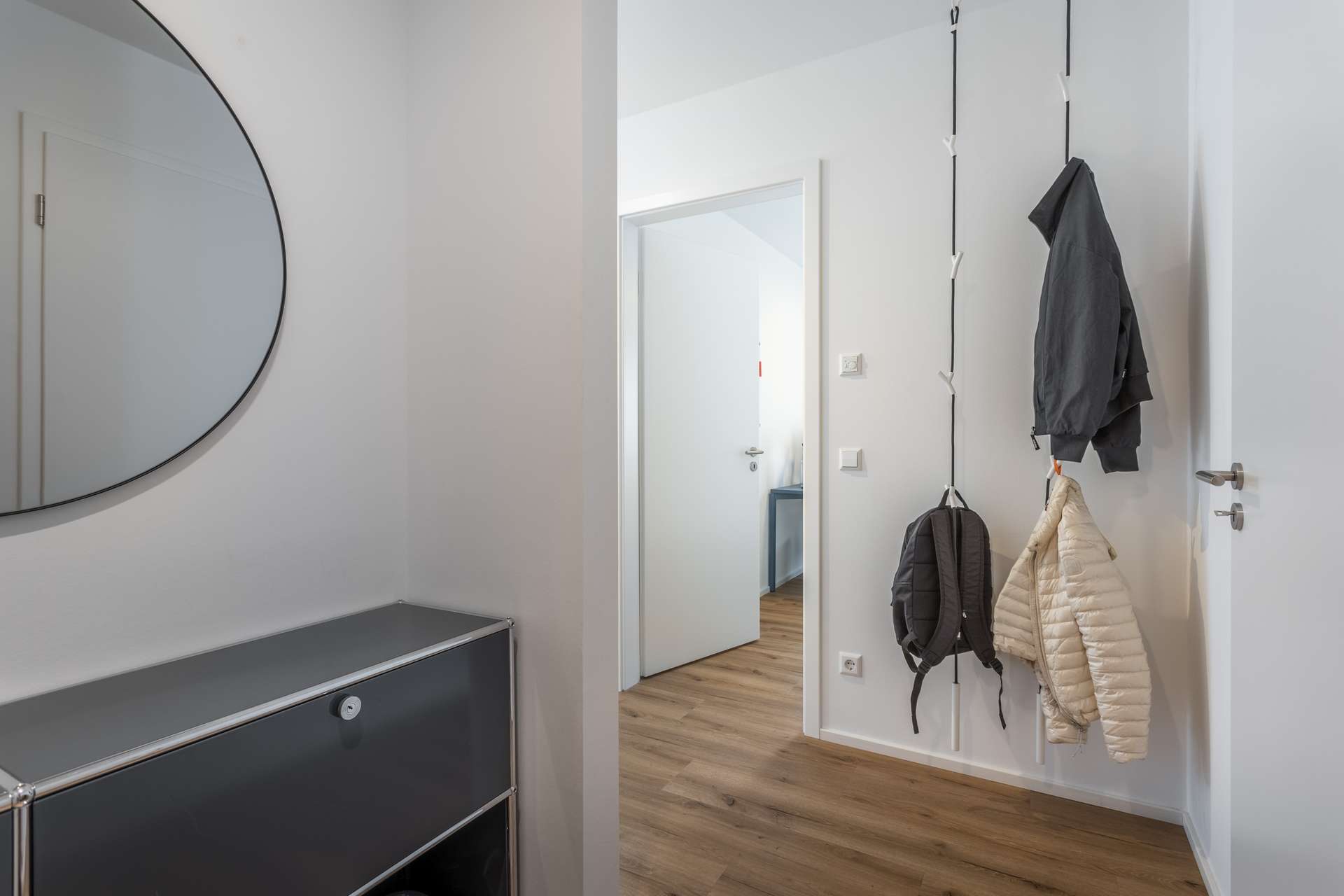 Flur Ein moderner Flur mit hellem Holzfußboden, weißen Wänden, einem runden Spiegel über einem schwarzen Schrank und zwei Jacken, die an Haken neben einer offenen Tür hängen, die zu einem anderen Raum führt.
