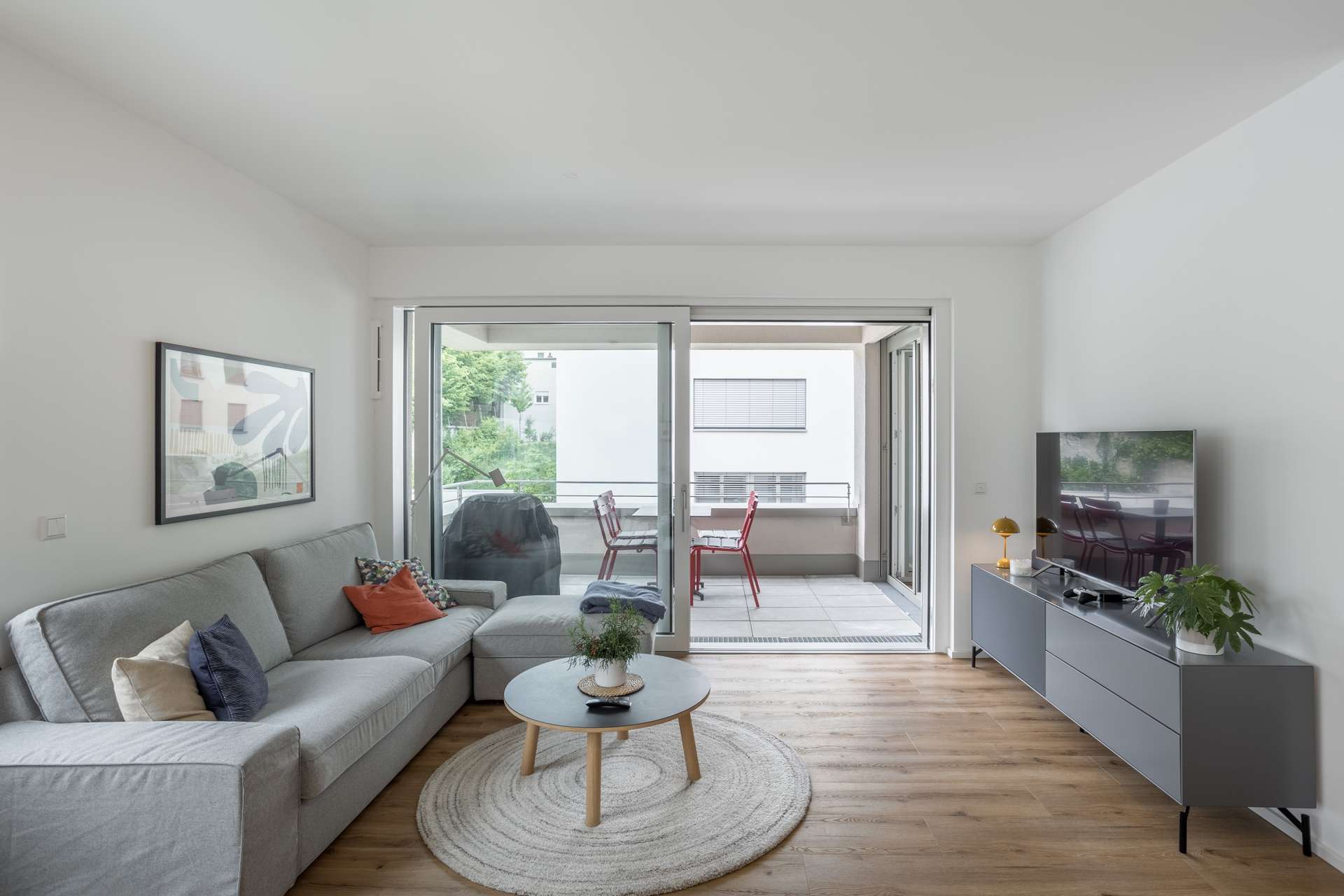 Wohn- und Essbereich Ein modernes Wohnzimmer mit einem grauen Sofa, einem runden Couchtisch, einem Fernseher auf einem Ständer und Wandkunst. Glastüren führen auf einen Balkon mit einem Tisch, roten Stühlen und Blick auf grüne Bäume und nahe gelegene Gebäude.