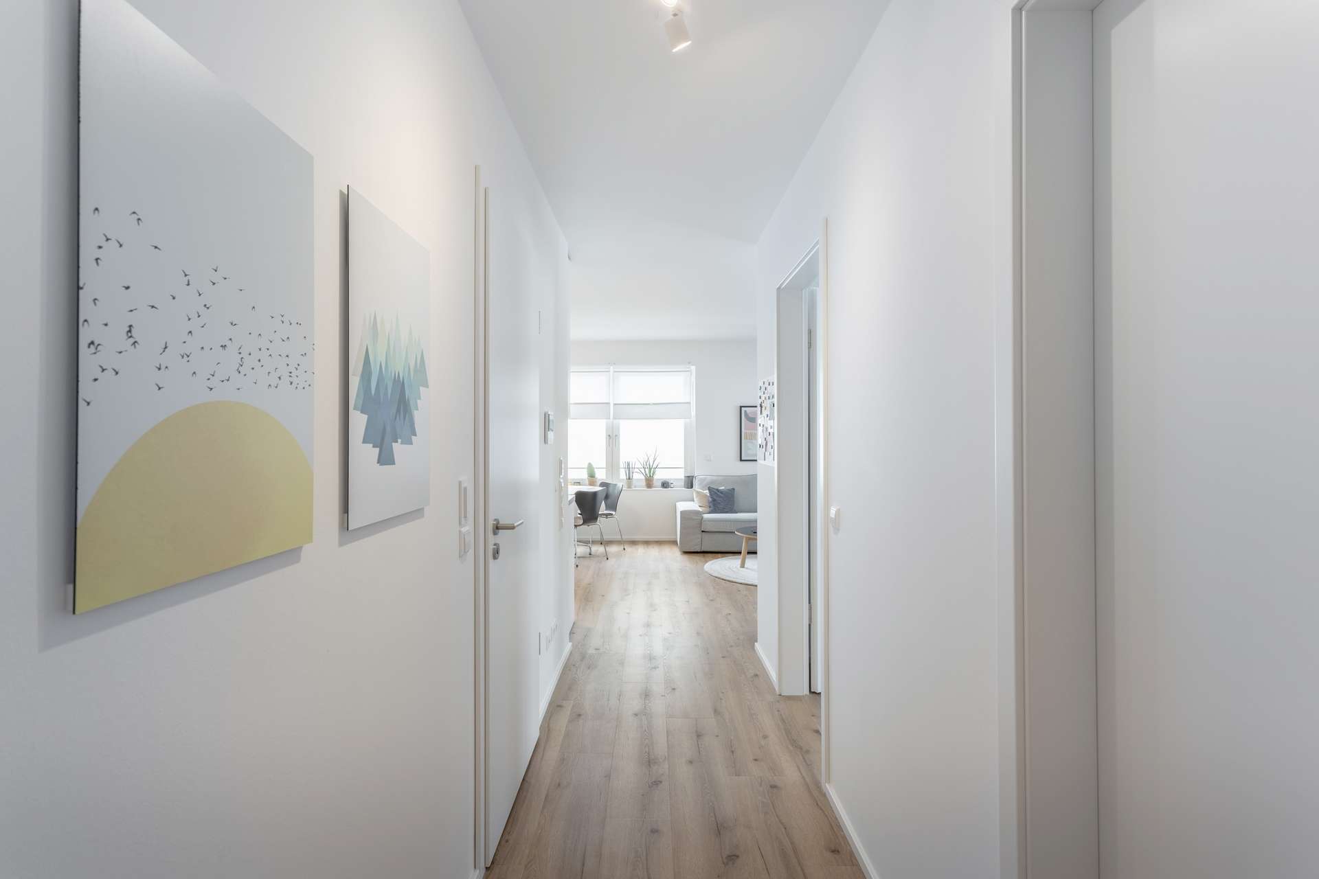Flur Ein heller, minimalistischer Flur mit hellem Holzfußboden, weißen Wänden und zwei gerahmten Kunstwerken. Der Flur führt zu einem Wohnzimmer mit großen Fenstern, die viel natürliches Licht hereinlassen. Auf beiden Seiten des Flurs sind Türen zu sehen.