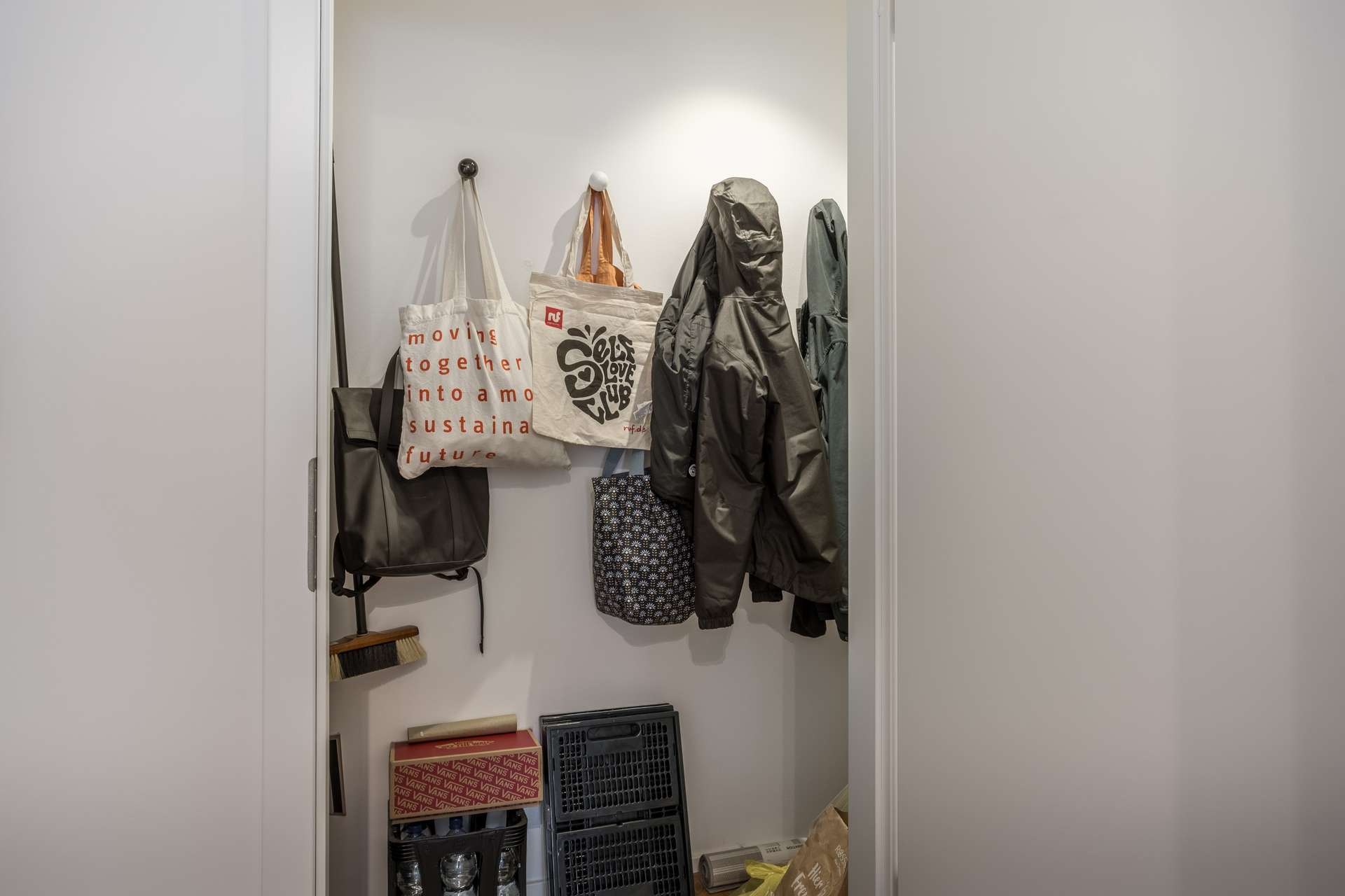 Abstellkammer Ein kleiner Schrank mit Jacken, Taschen und Behältern, die an Haken hängen. Auf dem Boden stehen Kisten, gefaltete Kartons und verschiedene Gegenstände, die in der Ecke gelagert werden. Der Raum ist hell erleuchtet, die Wände sind weiß.