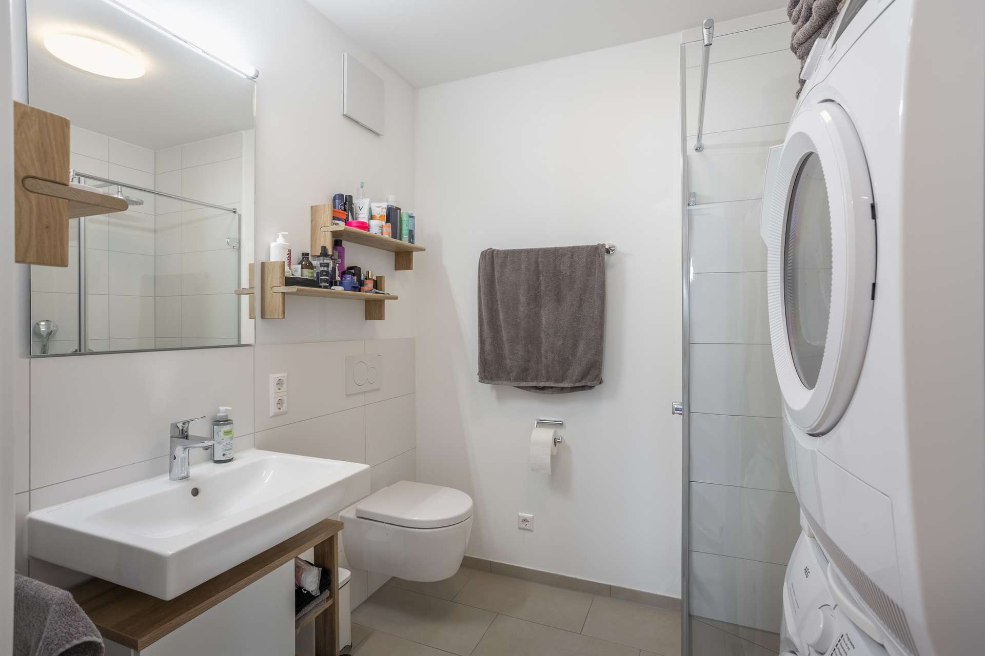 Badezimmer Modernes Badezimmer mit weißen Wänden, einem Waschbecken mit Spiegel, Regalen für Toilettenartikel, einer an der Wand befestigten Toilette, einem grauen Handtuch, einem Toilettenpapierhalter und einer gestapelten Waschmaschine und einem Trockner neben einer Glasduschkabine.